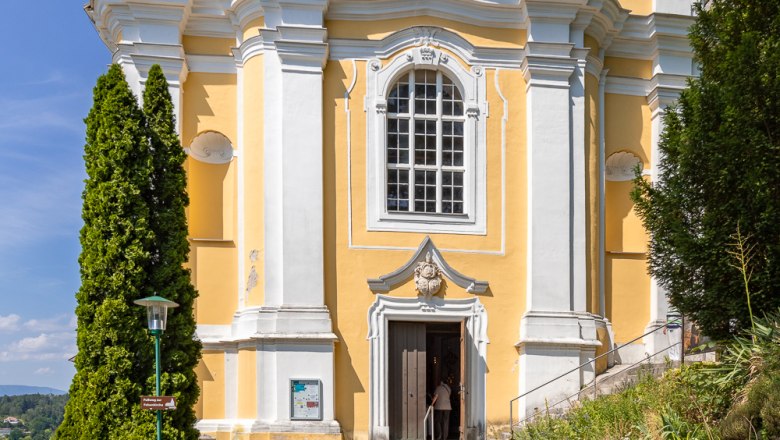 Gelbe Barockkirche mit weißen Säulen und einer Statue über dem Eingang, umgeben von Bäumen und blauem Himmel.