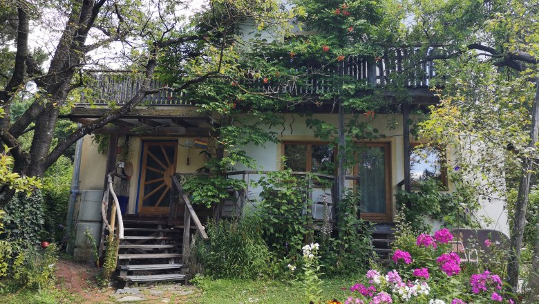 Ein Haus mit Holzbalkon, umgeben von Bäumen und bunten Blumen im Garten.