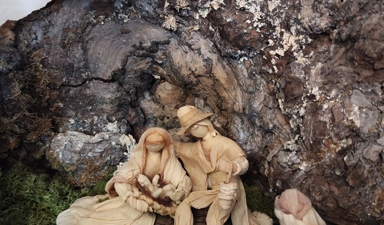 Naturkrippe mit Figuren aus Maisschalen unter einem Baumstamm.