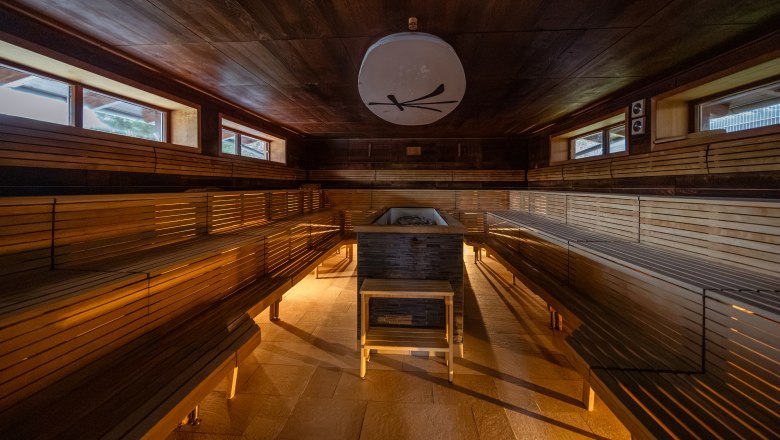 Innenansicht einer geräumigen Sauna mit Holzbänken und einem Ofen in der Mitte.