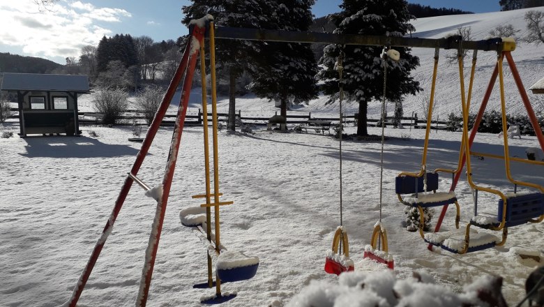 Schneebedeckter Spielplatz mit Schaukeln und B&auml;umen im Hintergrund.