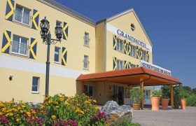 Au&szlig;enansicht eines gelben Hotels mit blauen Fensterl&auml;den und einem Vordach. Vor dem Geb&auml;ude bl&uuml;hen gelbe und pinke Blumen.