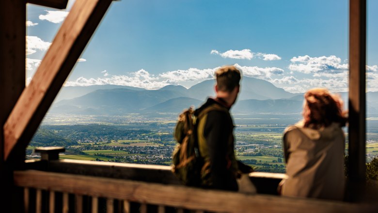 Zwei Personen genießen die Aussicht von einem Aussichtspunkt auf eine weite Landschaft mit Bergen im Hintergrund.