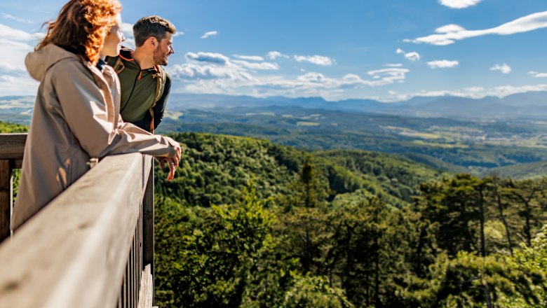 Zwei Personen stehen auf der Aussichtswarte und blicken über eine bewaldete, hügelige Landschaft.