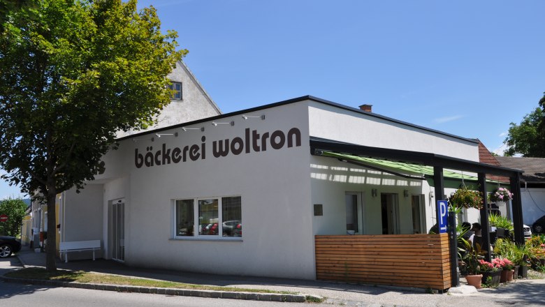 Außenansicht der Bäckerei Woltron mit Pflanzen und Parkplätzen.