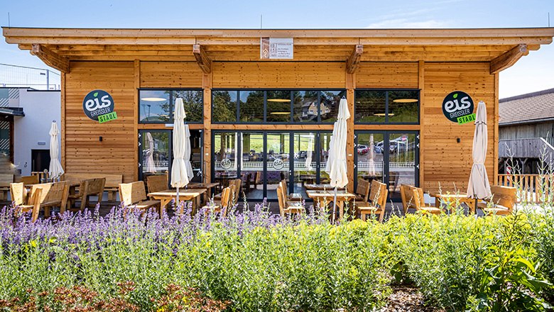 Modernes Holzgebäude mit Terasse, auf der Bänke, Stühle und Sonnenschirme stehen.