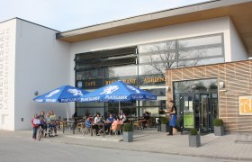 Außenansicht des Cafe Adrienn mit Gästen unter blauen Sonnenschirmen.