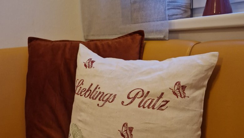 Ein gem&uuml;tliches Sofa mit einem dekorativen Kissen, auf dem 'Lieblings Platz' steht, neben einem Fenster mit Pflanzen und einer Vase.