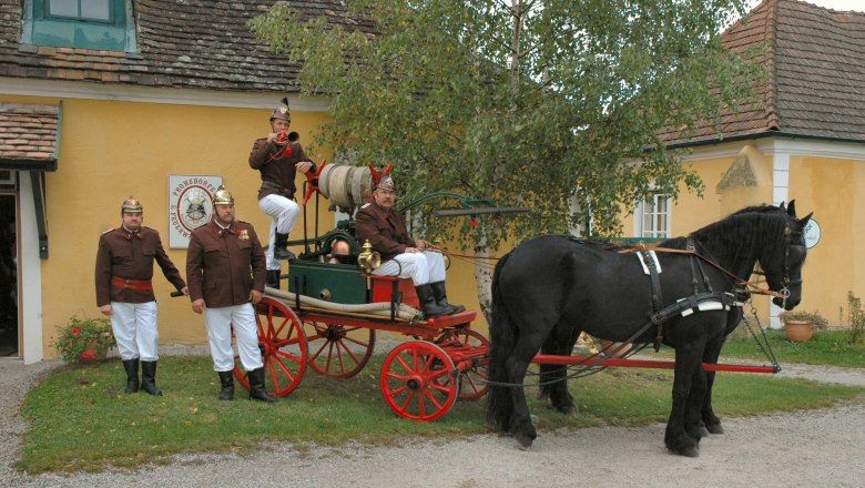 Historische Feuerwehrkutsche mit Pferd und Feuerwehrleuten in Uniform vor einem gelben Gebäude.