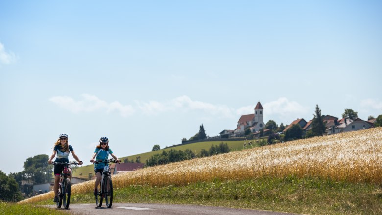 Zwei Personen fahren mit E-Bikes auf einem l&auml;ndlichen Weg, im Hintergrund ein Dorf mit Kirche.