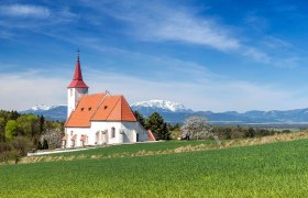 Wei&szlig;e Pfarrkirche Ofenbach mit rotem Dach und Kirchturm vor einer Bergkulisse mit gr&uuml;nem Feld im Vordergrund.