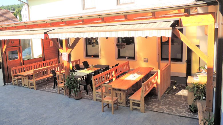 Ein gem&uuml;tlicher Gastgarten mit Holzb&auml;nken und Tischen unter einer Markise.