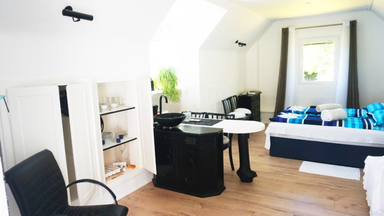 Innenansicht eines hellen Ferienhauses mit Bett, Tisch, St&uuml;hlen und K&uuml;chenzeile.