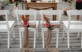 Weiße Stühle mit Blumen und Stoff dekoriert auf einer Holzterrasse, vorbereitet für eine Hochzeit.