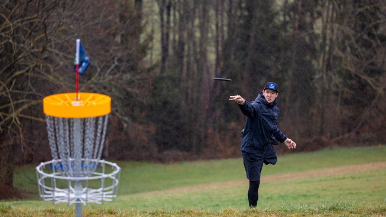 Ein Mann wirft eine Disc in Richtung eines Discgolf-Korbs auf einer Wiese, umgeben von Bäumen.
