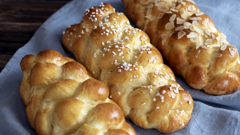 Drei geflochtene Brioche-Striezel auf einem blauen Tuch, einer mit Mandeln, einer mit Hagelzucker bestreut.