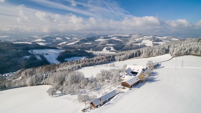 Verschneite Landschaft mit Bauernhof und Hügeln im Hintergrund.