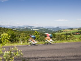Zwei Personen am Rennrad fahren schnell auf einer Stra&szlig;e in einer h&uuml;geligen Landschaft.