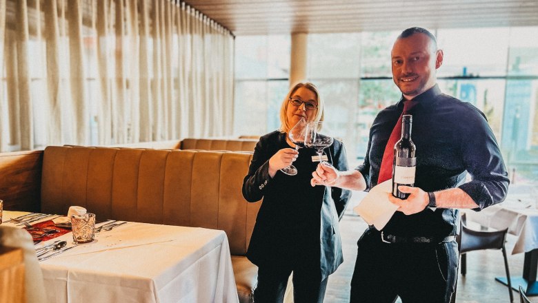 Zwei Personen in einem Restaurant stoßen mit Weingläsern an, eine Person hält eine Weinflasche.