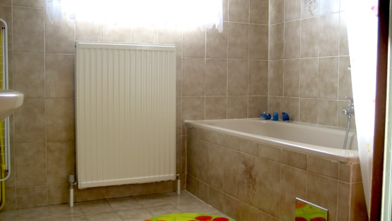 Ein Badezimmer mit beiger Fliesenwand, Badewanne, Heizk&ouml;rper und bunten Teppichen.