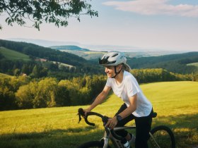 Eine Person f&auml;hrt mit einem Fahrrad durch eine h&uuml;gelige Landschaft mit gr&uuml;nen Wiesen und W&auml;ldern im Hintergrund.