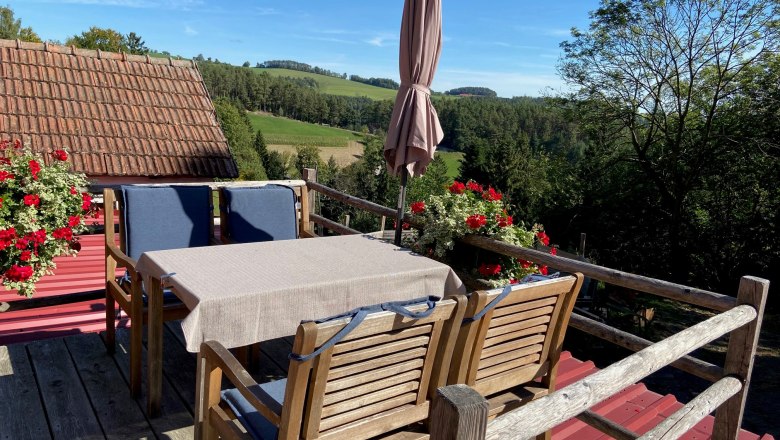 Kleine Terrasse mit Holzmöbeln, Sonnenschirm und Blumenkästen, umgeben von grüner Landschaft.