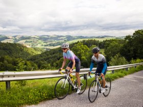 Zwei Radfahrende fahren auf einer Stra&szlig;e in einer h&uuml;geligen Landschaft.