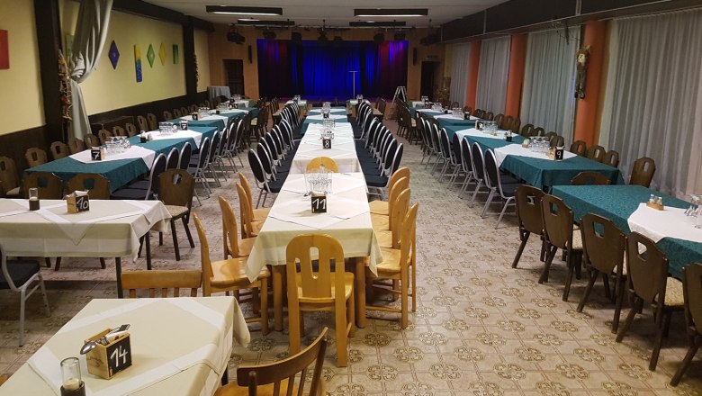 Ein leerer Veranstaltungssaal mit gedeckten Tischen und einer B&uuml;hne im Hintergrund.