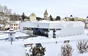 Modernes Geb&auml;ude im Schnee mit Kirche im Hintergrund.