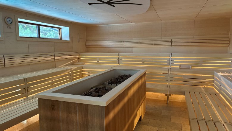 Innenansicht einer modernen Sauna mit Holzbänken und einem Ofen in der Mitte.