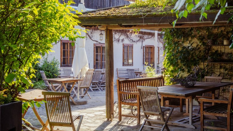 Ein sonniger Garten mit Holzmöbeln und einem überdachten Sitzbereich, umgeben von Pflanzen und einem Gebäude im Hintergrund.