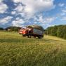 Ein Traktor mit Anhänger voller Heu fährt über ein grünes Feld unter einem blauen Himmel mit Wolken.