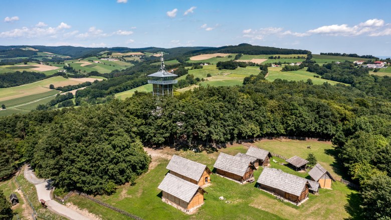 Luftaufnahme des Keltendorfs Schwarzenbach mit Holzh&auml;usern und einem Aussichtsturm, umgeben von gr&uuml;nen Feldern und W&auml;ldern.