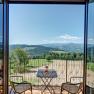 Blick von einem Balkon auf eine hügelige Landschaft mit Wäldern und Feldern, zwei Stühle und ein Tisch mit Blumen im Vordergrund.