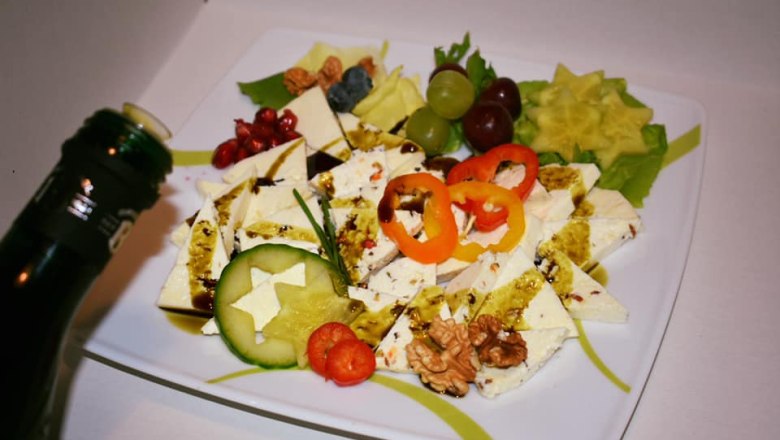 Ein Teller mit Schafkäse, dekoriert mit Paprika, Gurke, Nüssen und Früchten. Eine Flasche mit Öl wird darüber gehalten.