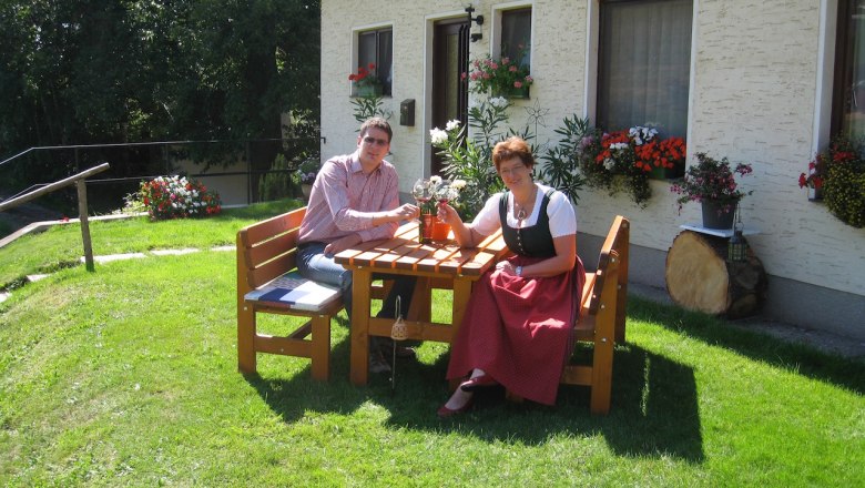 Zwei Personen sitzen an einem Holztisch im Garten vor einem Haus mit Blumen.