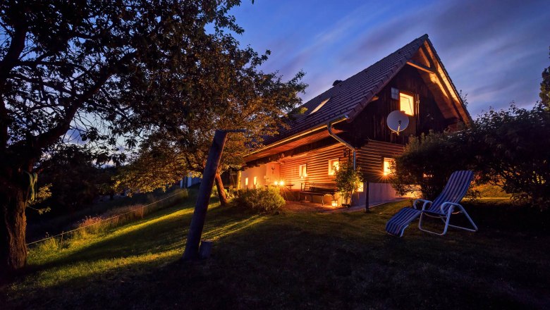 Ein beleuchtetes Ferienhaus bei Nacht mit Garten und Liegestuhl.