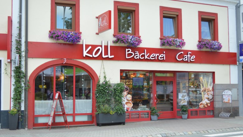 Au&szlig;enansicht der Koll B&auml;ckerei Caf&eacute; in Kirchschlag mit roten Fensterrahmen und Blumenk&auml;sten.