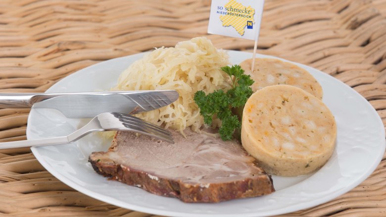 Weißer Teller auf einem Korbtisch mit Schweinebraten, Knödeln und Sauerkraut, garniert mit Petersilie.
