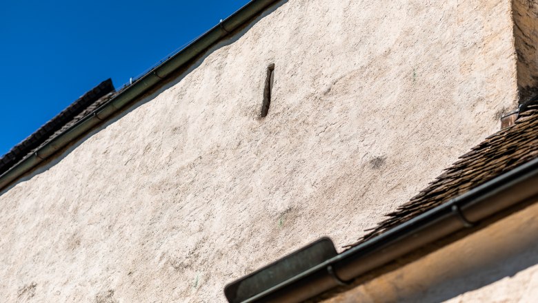Nahaufnahme einer wei&szlig;en Mauer mit Schie&szlig;scharte Dachrinne und blauen Himmel im Hintergrund.