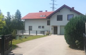 Zweist&ouml;ckiges Haus mit rotem Dach und Garage, umgeben von B&auml;umen und Str&auml;uchern.