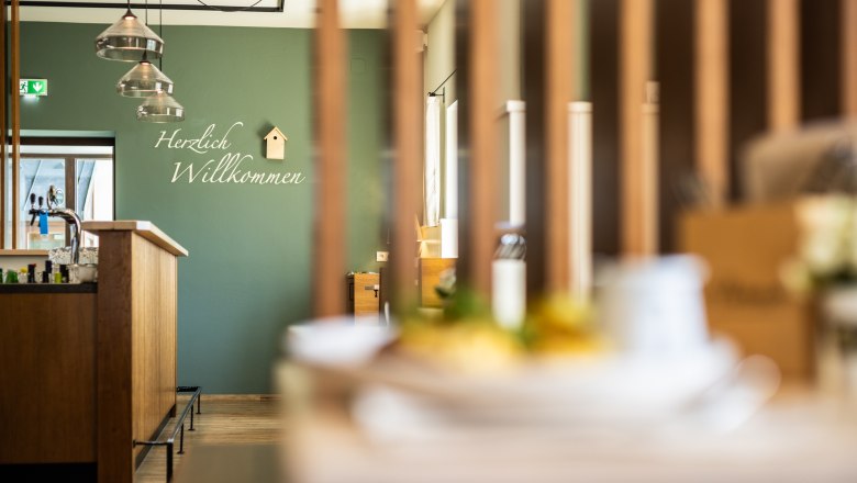 Innenansicht eines modernen Gastraumes mit grüner Wand und "Herzlich Willkommen"-Schriftzug. Über der Schank aus Holz hängen Lampen und an der Wand ein kleines Vogelhaus zur Verzierung.
