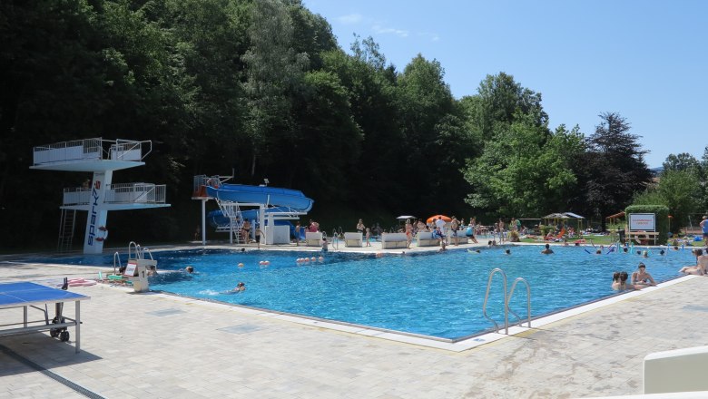 Ein Freibad mit Sprungturm, Rutsche und vielen Badeg&auml;sten an einem sonnigen Tag.