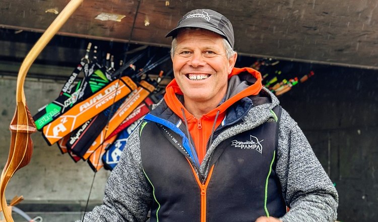 Ein l&auml;chelnder Mann h&auml;lt einen Bogen vor einem Anh&auml;nger mit Pfeilen.