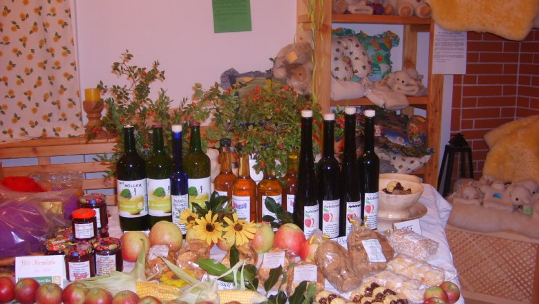 Ein Tisch in einem Hofladen mit verschiedenen Bio-Produkten wie &Auml;pfeln, Marmeladen, S&auml;ften und Lik&ouml;ren, dekoriert mit Blumen und Pflanzen.