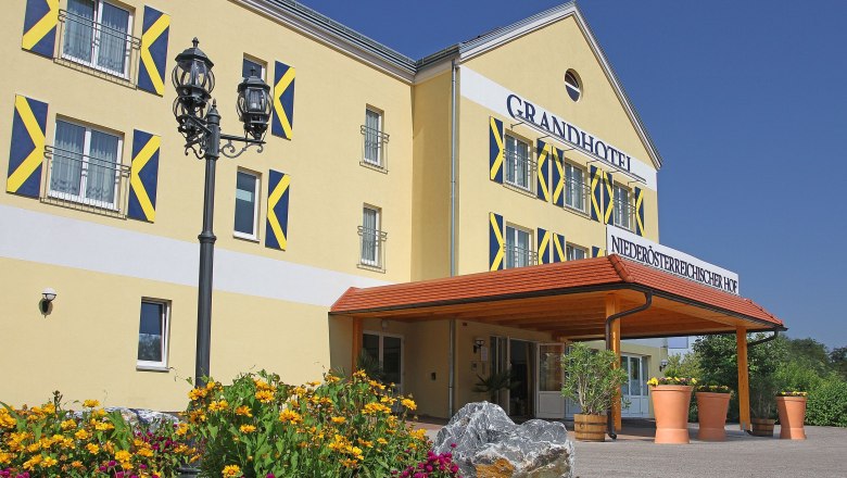 Au&szlig;enansicht eines gelben Hotels mit blauen Fensterl&auml;den und einem Vordach. Vor dem Geb&auml;ude bl&uuml;hen gelbe und pinke Blumen.