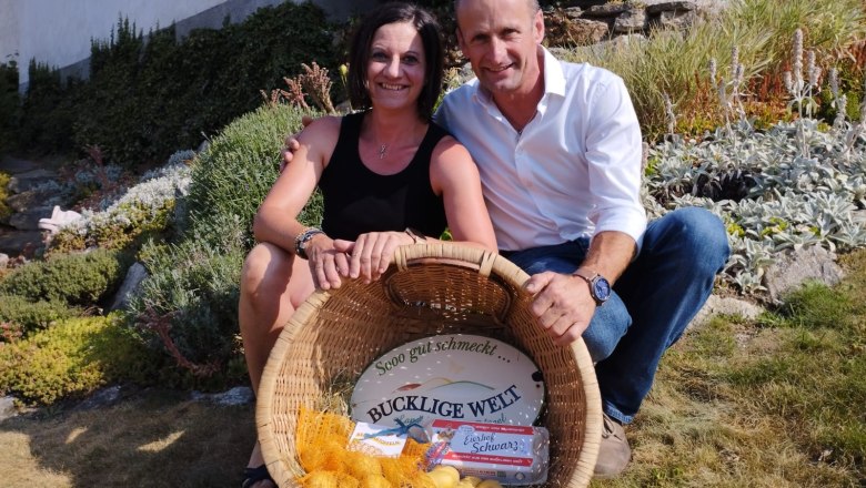 Eine Frau und ein Mann sitzen l&auml;chelnd in einem Garten und halten einen Korb mit Kartoffeln und Eiern.