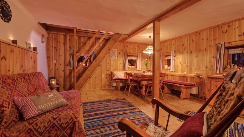 Gem&uuml;tliches Wohnzimmer mit Holzw&auml;nden, Sofa, Schaukelstuhl und Esstisch. Rustikales Ambiente mit Teppich und warmem Licht.