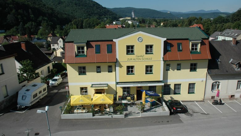 Gelbes Gebäude mit der Aufschrift 'Gasthof Zur Alten Schule', umgeben von Hügeln und Bäumen.