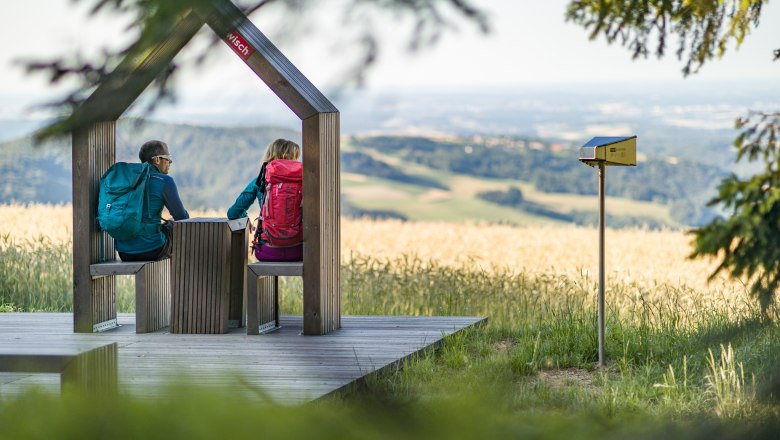Zwei Wandernde sitzen auf einer Holzplattform mit Aussicht auf eine hügelige Landschaft.